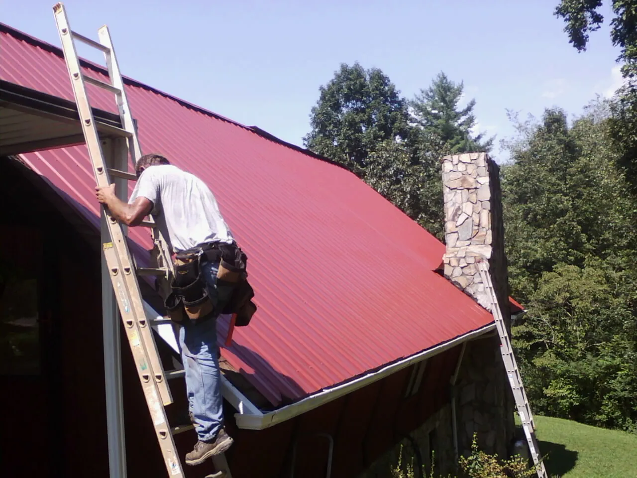 Roofer on a ladder beside a red metal roof and stone chimney — Soffit & Fascia Repair by Roofing Brothers in Fort Branch